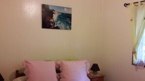Une chambre à coucher avec un lit, deux oreillers, une table de chevet avec une lampe, un tableau représentant un paysage côtier et une fenêtre avec des rideaux.