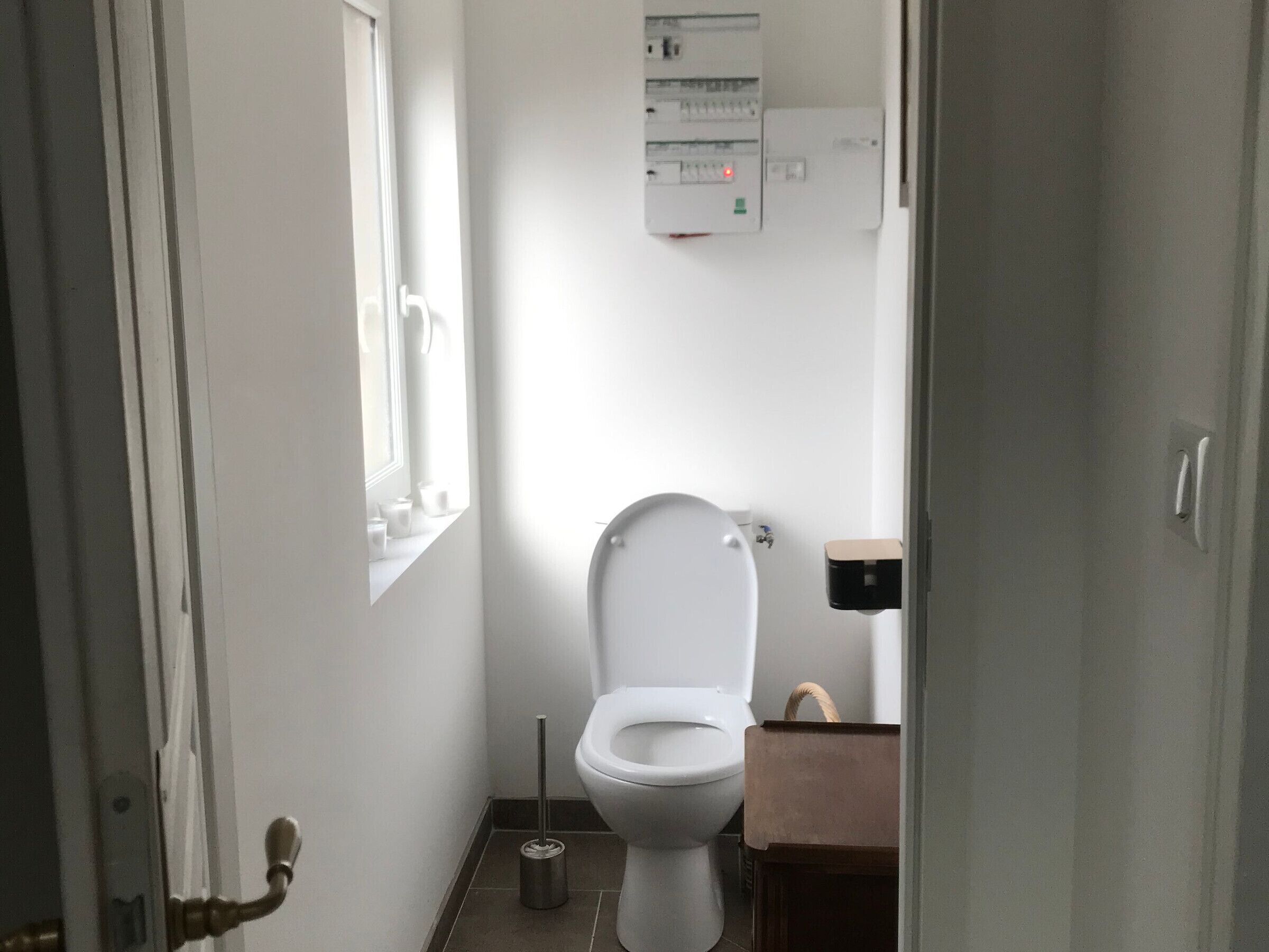 A small, clean bathroom with a toilet, a window, and a wall-mounted electrical panel.
