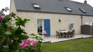A stone building with a grey roof, a blue door, and a patio with a table and chairs.