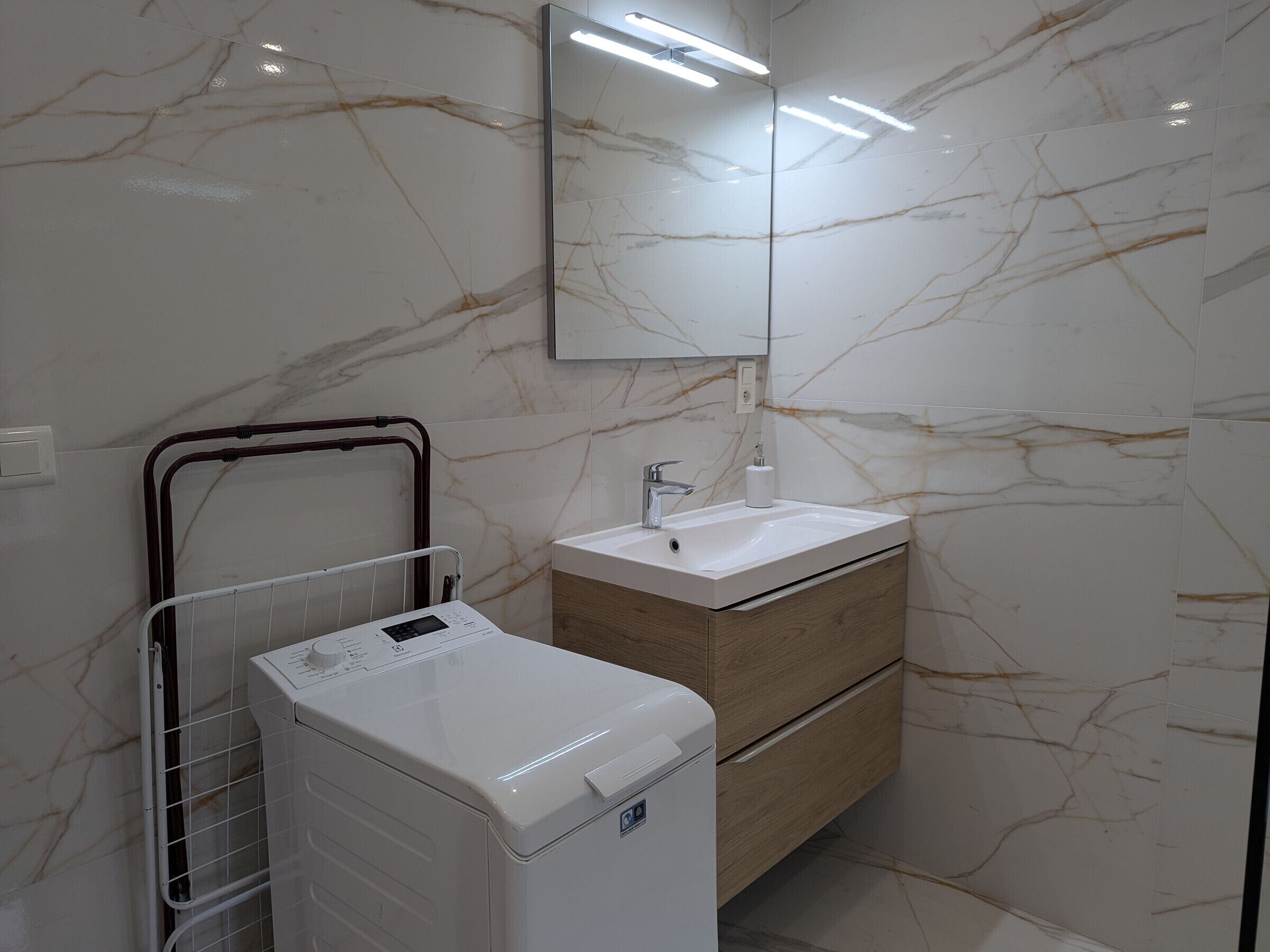 Une salle de bain moderne avec un pare-feu en marbre, un lave-linge blanc, un meuble-lavabo en bois et un miroir au-dessus du lavabo.