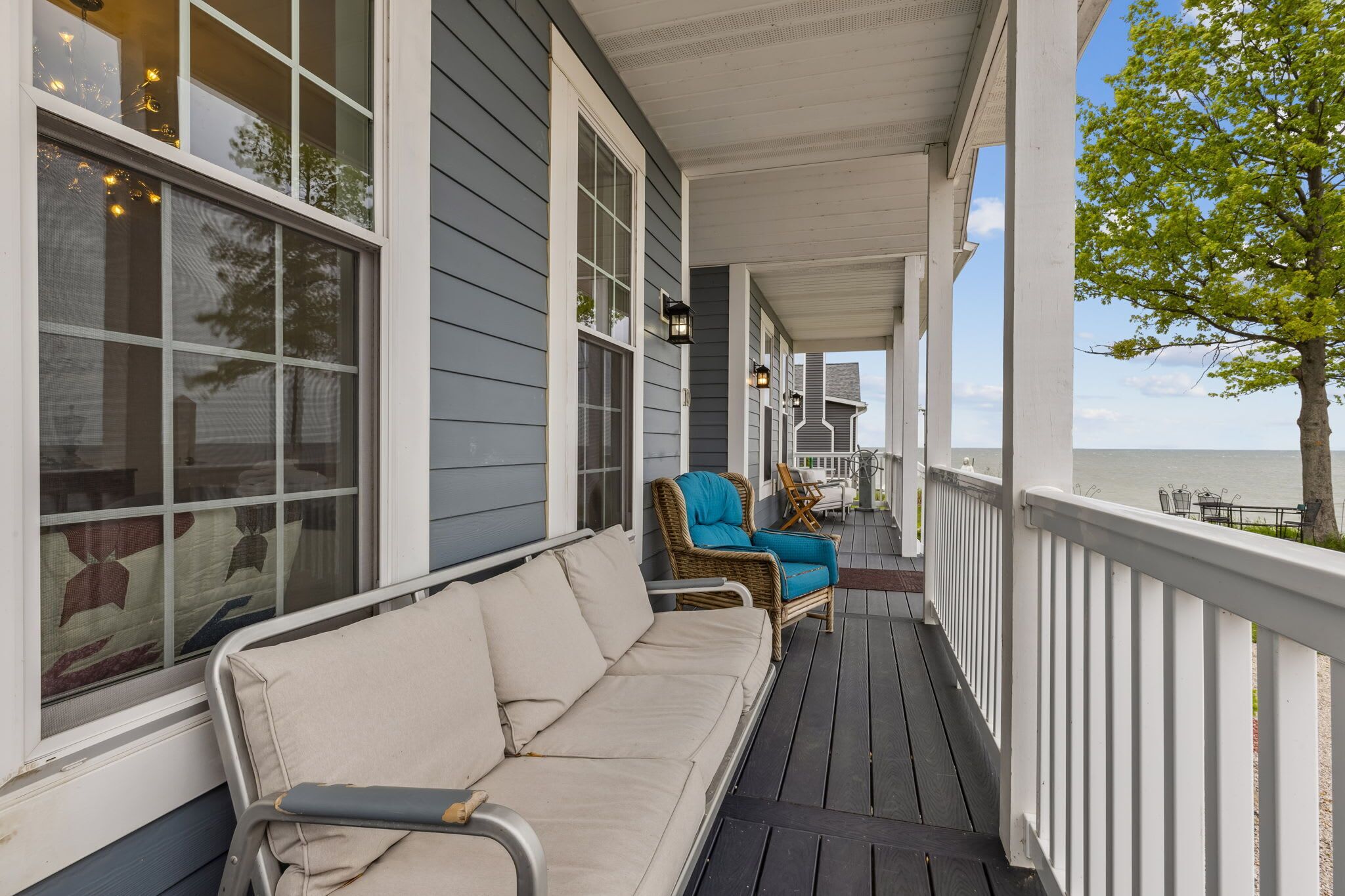 A covered porch with a beige sofa and two blue chairs, overlooking a body of water.