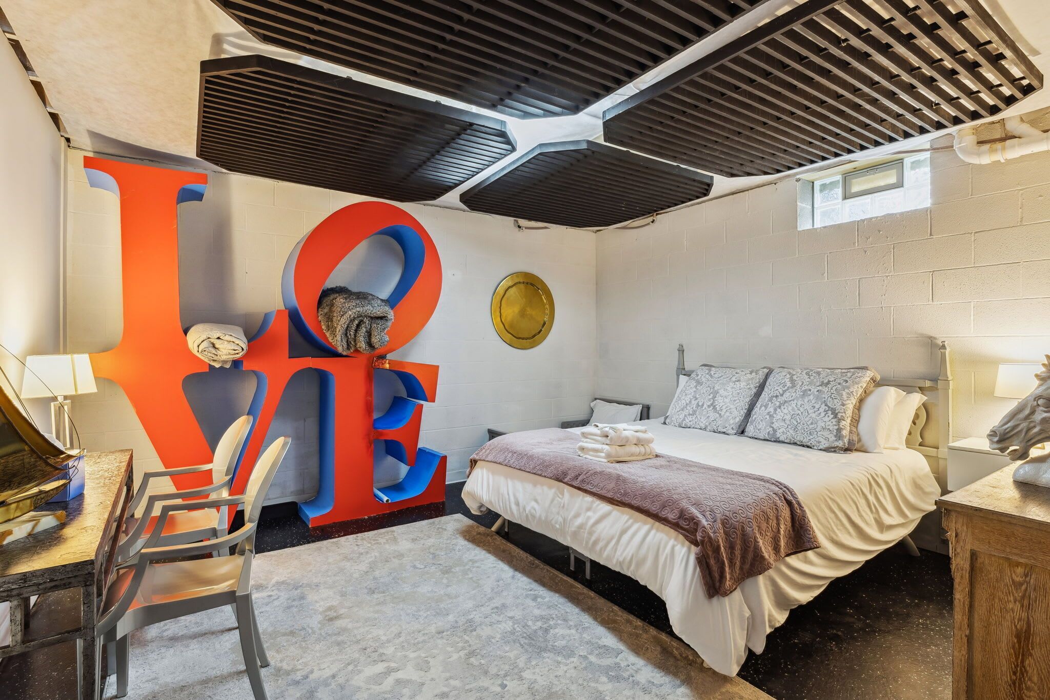 A modern bedroom with a large 'LOVE' wall art, a bed with a patterned blanket, a desk with a lamp, and a chair.