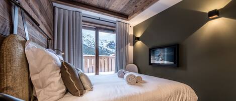Ein Schlafzimmer mit einem großen Bett, Holzbalkendecke, Bergblick durch das Fenster und einem an der Wand befestigten Fernseher.