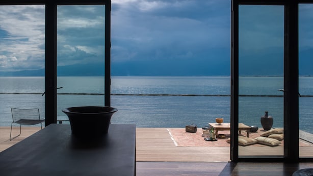 A room with a large window overlooking a body of water, a wooden table, a chair, and a small table with various items. - Yakamoz Erhai Hotel (Dali)