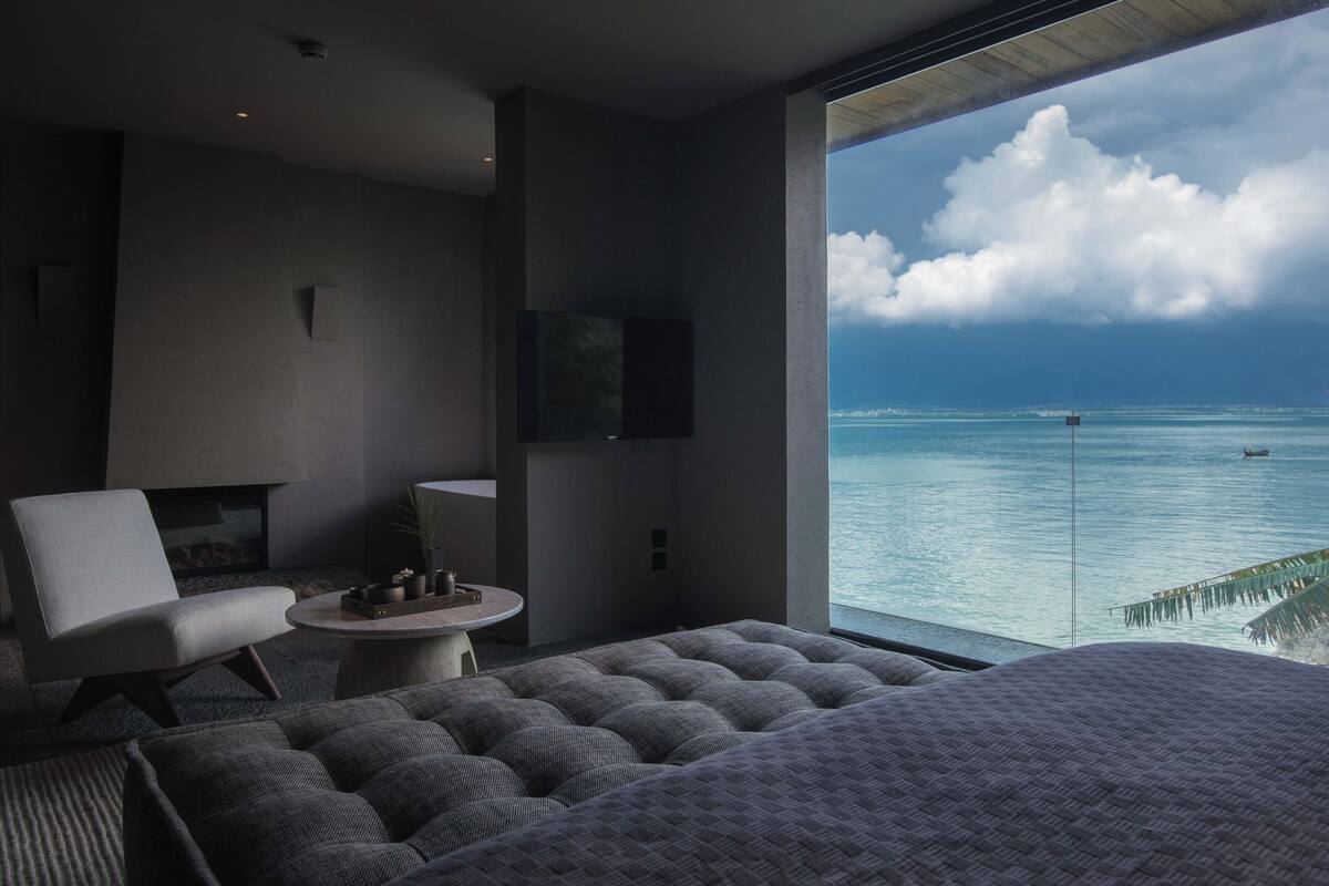 A modern bedroom with a large bed, a sofa, and a TV. It has a view of the ocean and a palm tree.