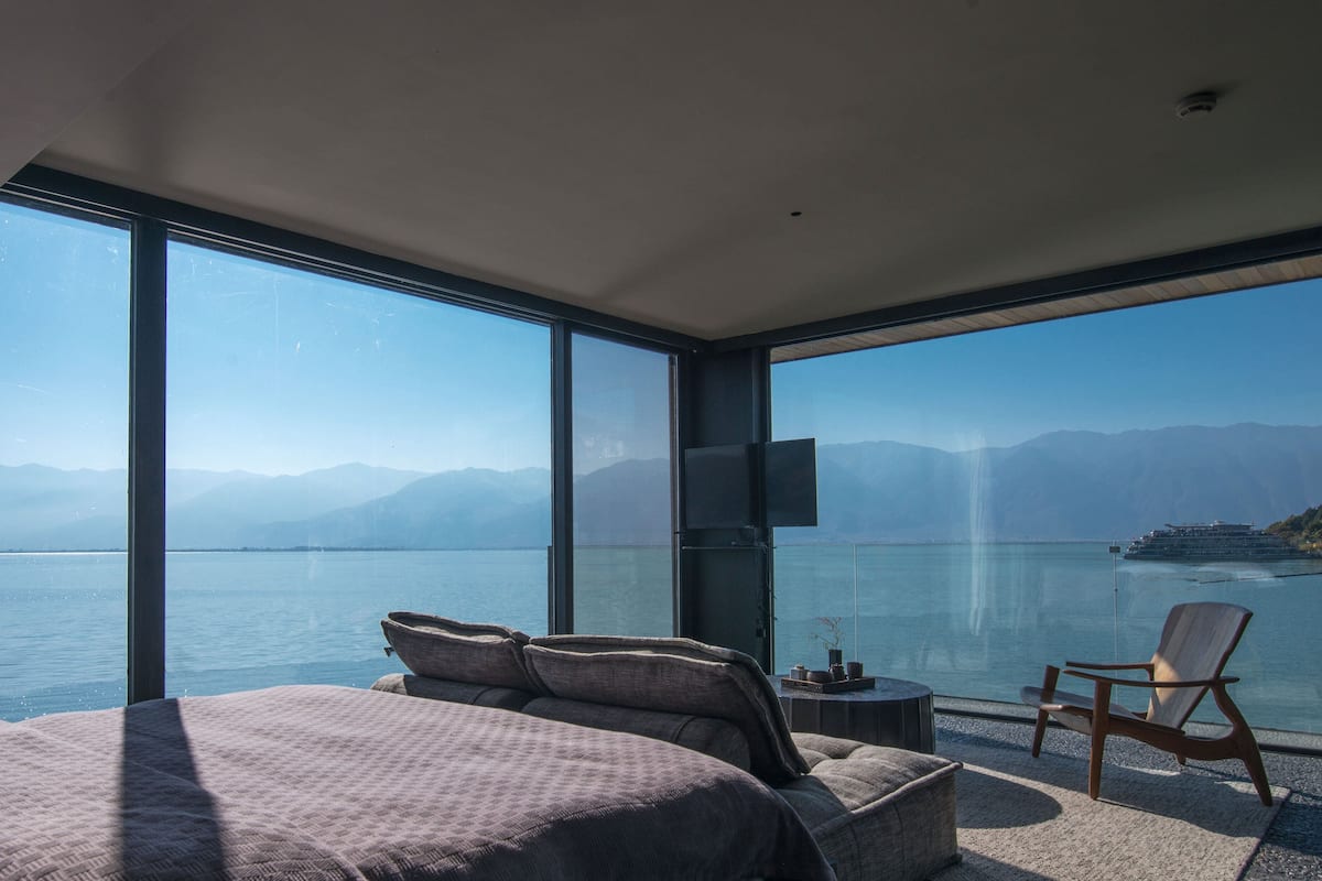 A modern bedroom with a large bed, a seating area with a chair, and a balcony overlooking a body of water and mountains.