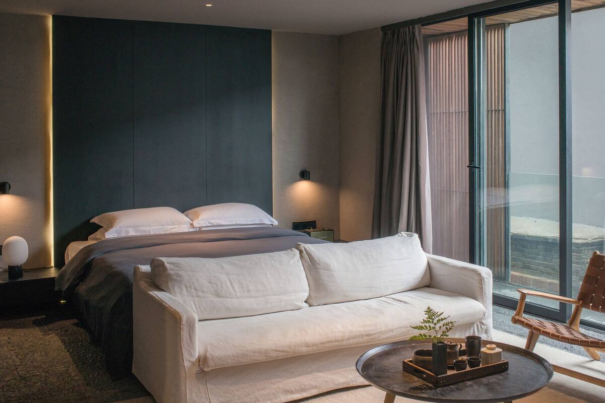 A modern hotel room with a large bed, a white sofa, a small round table, and a view of the city through large windows.
