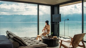A modern room with a large window overlooking a lake and mountains. There is a sofa, a wooden chair, and a small table with a hat and a plant. - Yakamoz Erhai Hotel (Dali)
