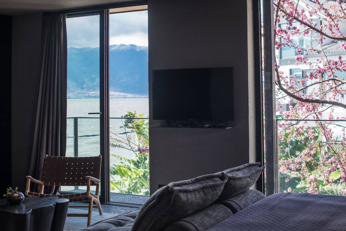 A hotel room with a large window offering a view of mountains and a body of water. There is a flat-screen TV mounted on the wall and a wooden chair by the window.