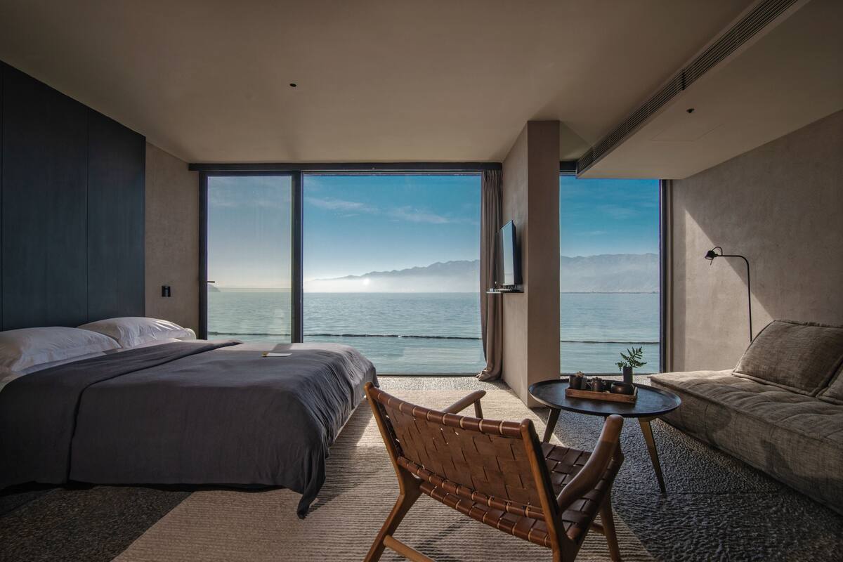 A modern hotel room with a large bed, a seating area with a table, and a view of the sea.