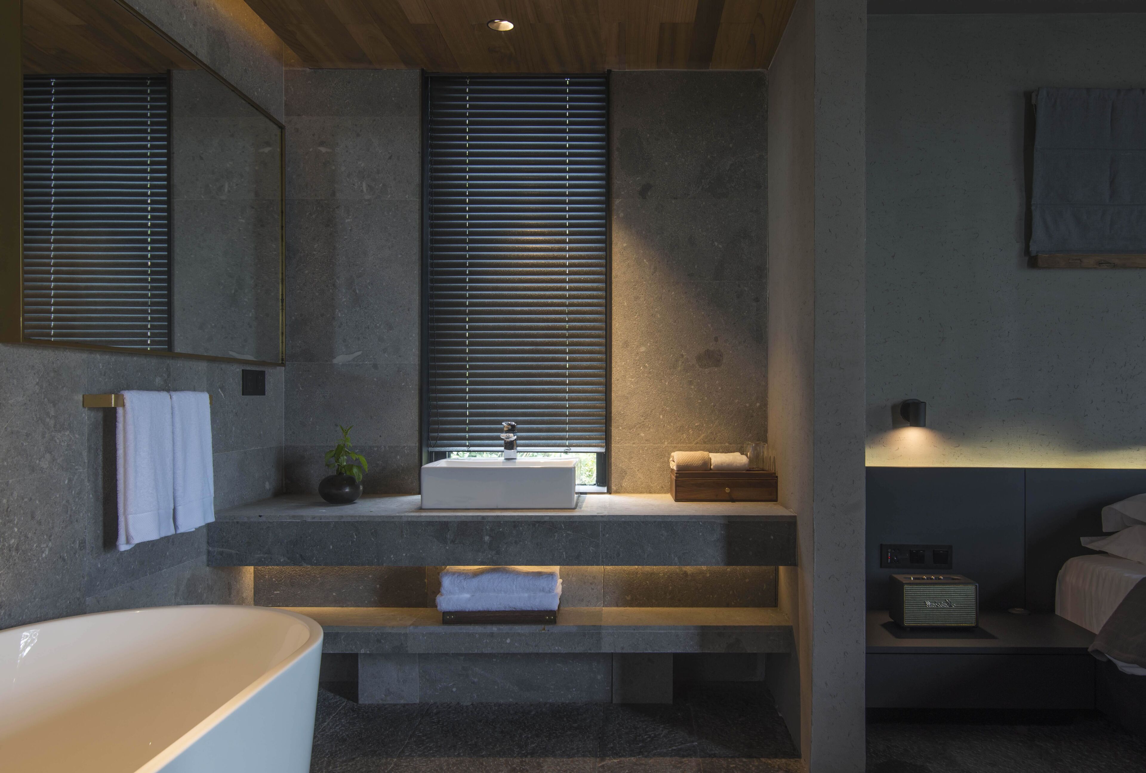 A modern bathroom with a freestanding bathtub, a sink with a window above, and a bedside table with a lamp.