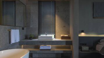A modern bathroom with a freestanding bathtub, a sink with a window above, and a bedside table with a lamp.