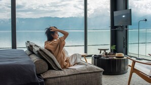 A room with a large window overlooking a body of water and mountains. There is a bed, a sofa, a TV, and a small table with a hat and a plant. - Yakamoz Erhai Hotel (Dali)