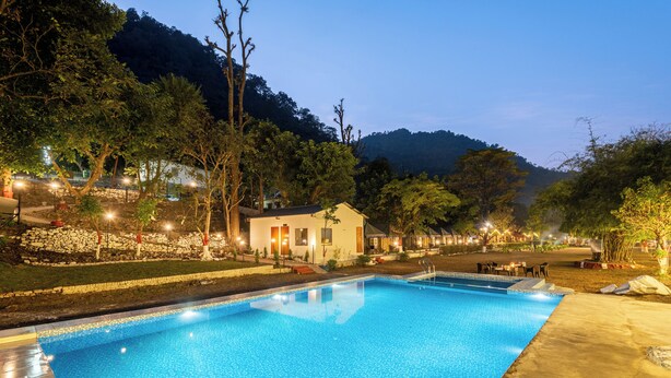 A swimming pool with a clear blue water surface, surrounded by trees and a mountain in the background. - Royal River Resort Rishikesh  (Rishikesh)