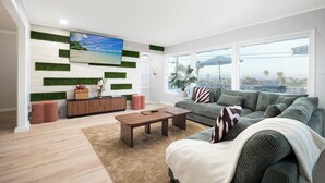 Una sala moderna con un televisor de pantalla plana grande, un sofá verde, una mesa de centro de madera y vistas al exterior a través de amplios ventanales.