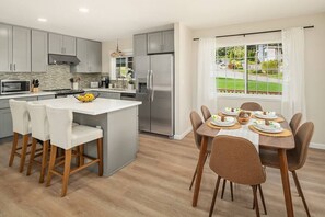 Una cucina moderna con mobili grigi, un'isola centrale con sgabelli, un frigorifero in acciaio inossidabile e una zona pranzo con tavolo apparecchiato per sei.
