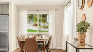 Un coin repas avec une table dressée pour quatre personnes, un réfrigérateur et une vue sur un jardin par la fenêtre.