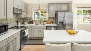 Une cuisine moderne avec des armoires grises, des appareils en acier inoxydable, un îlot central agrémenté d’un saladier de citrons et une vue sur le jardin à travers une fenêtre.