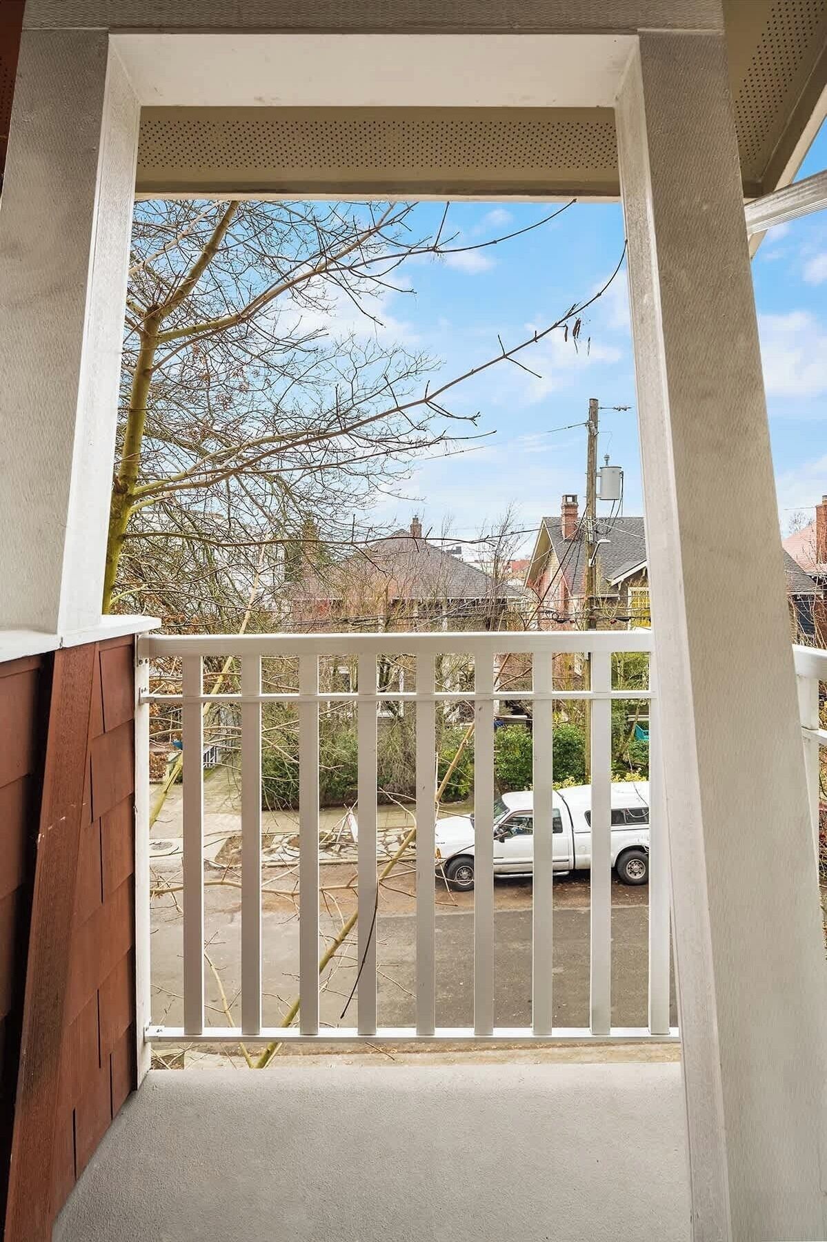 A balcony with a white railing, a view of a parked car, and leafless trees against a clear sky.