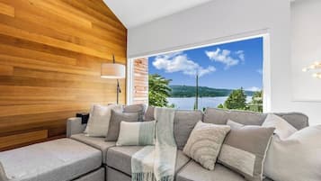 A modern living room with a sectional sofa, a wooden accent wall, and a large window offering a scenic view of a lake and mountains.
