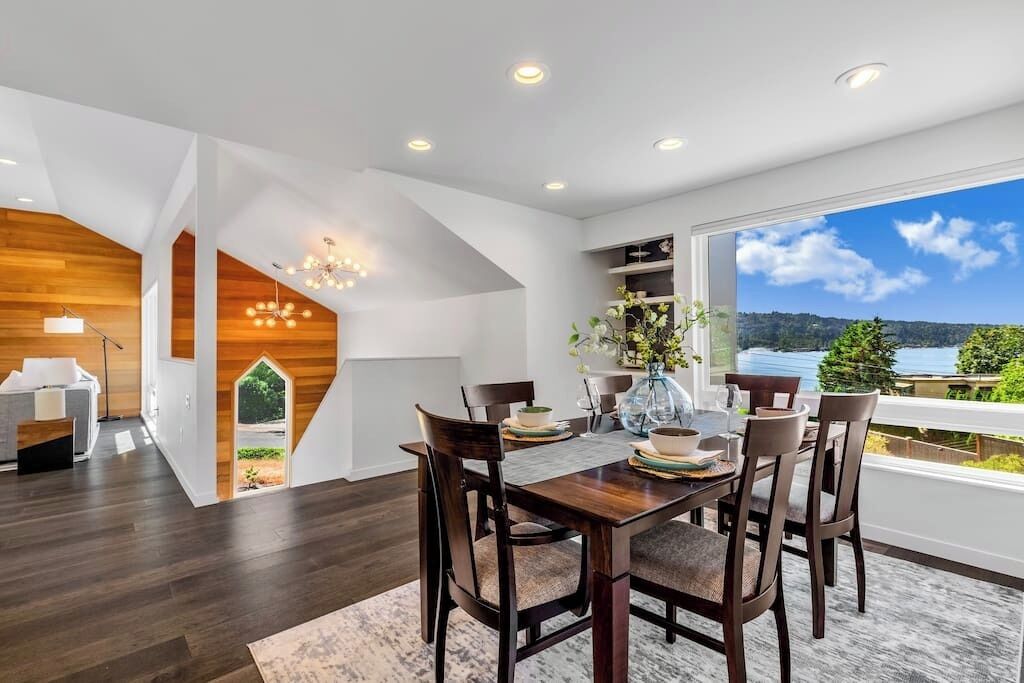 A modern dining area with a wooden table and chairs, a vase with flowers, and a view of a lake through large windows.