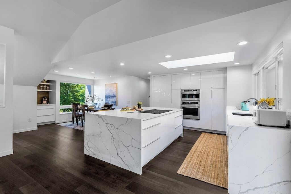 A modern kitchen with white cabinetry, a marble island, and a large window overlooking greenery.