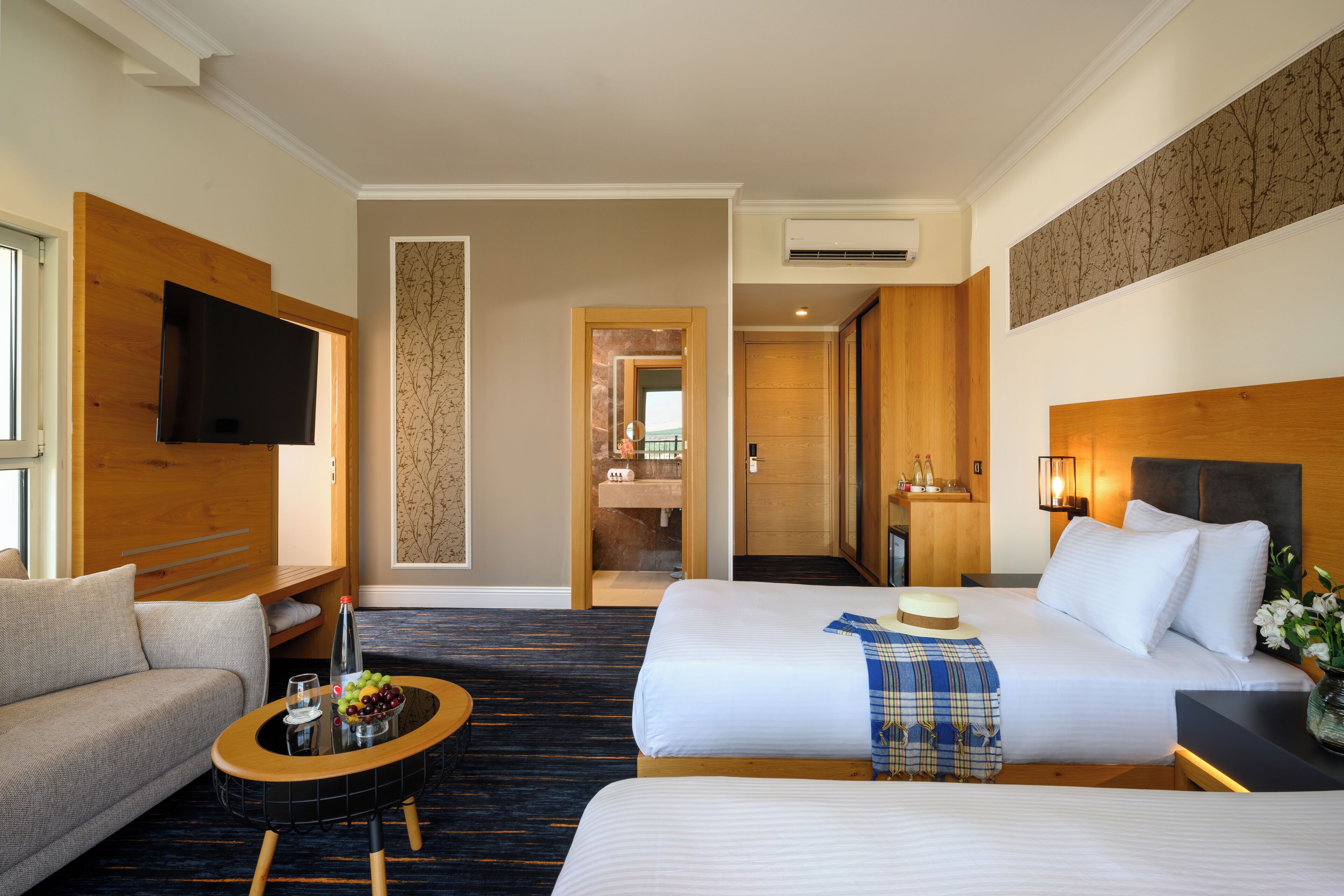 Habitación de hotel con cama, sofá, una mesa de centro con frutas y una botella, y un televisor fijado en la pared.