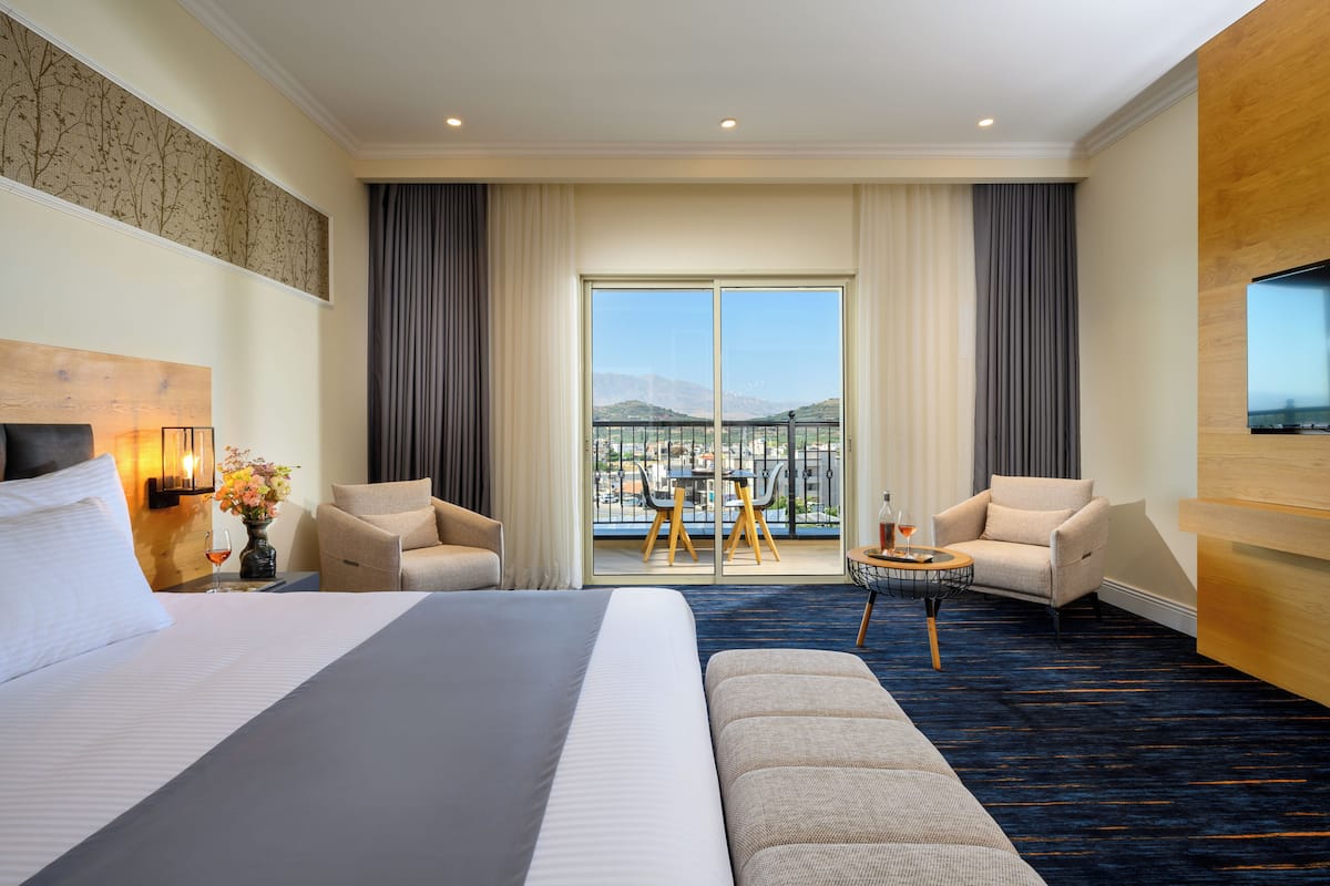 Ein Hotelzimmer mit einem großen Bett, zwei Sesseln, einem kleinen Tisch, einem Fernseher und einem Balkon mit Blick auf die Stadt und die Berge.