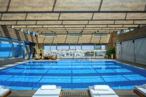 An indoor swimming pool with clear blue tiles, lounge chairs, and a view of the city. - OAKS HOTEL (Buq'ata)