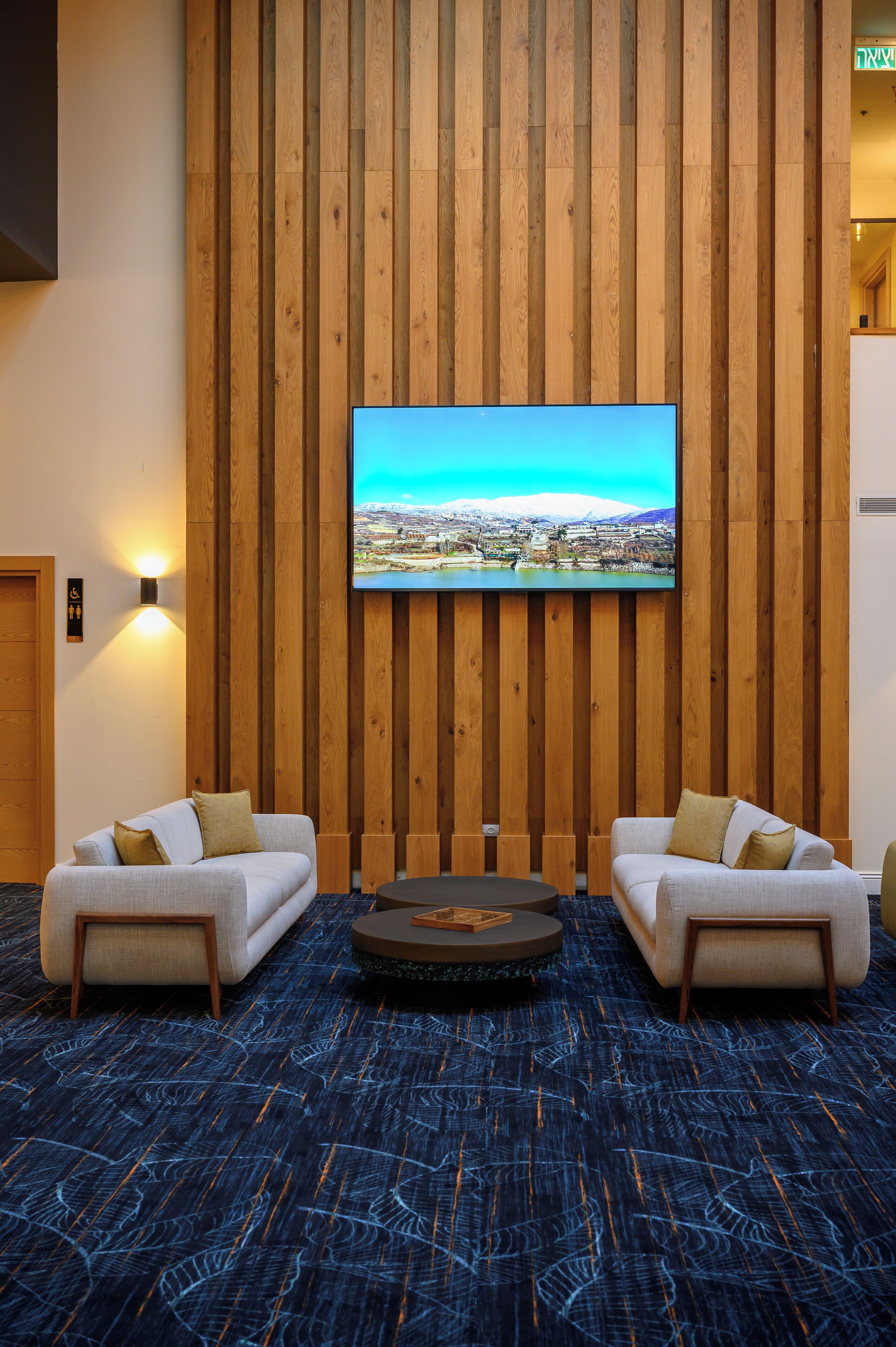 A room with a large TV mounted on a wooden wall, two sofas with cushions, a patterned carpet, and a small circular table.