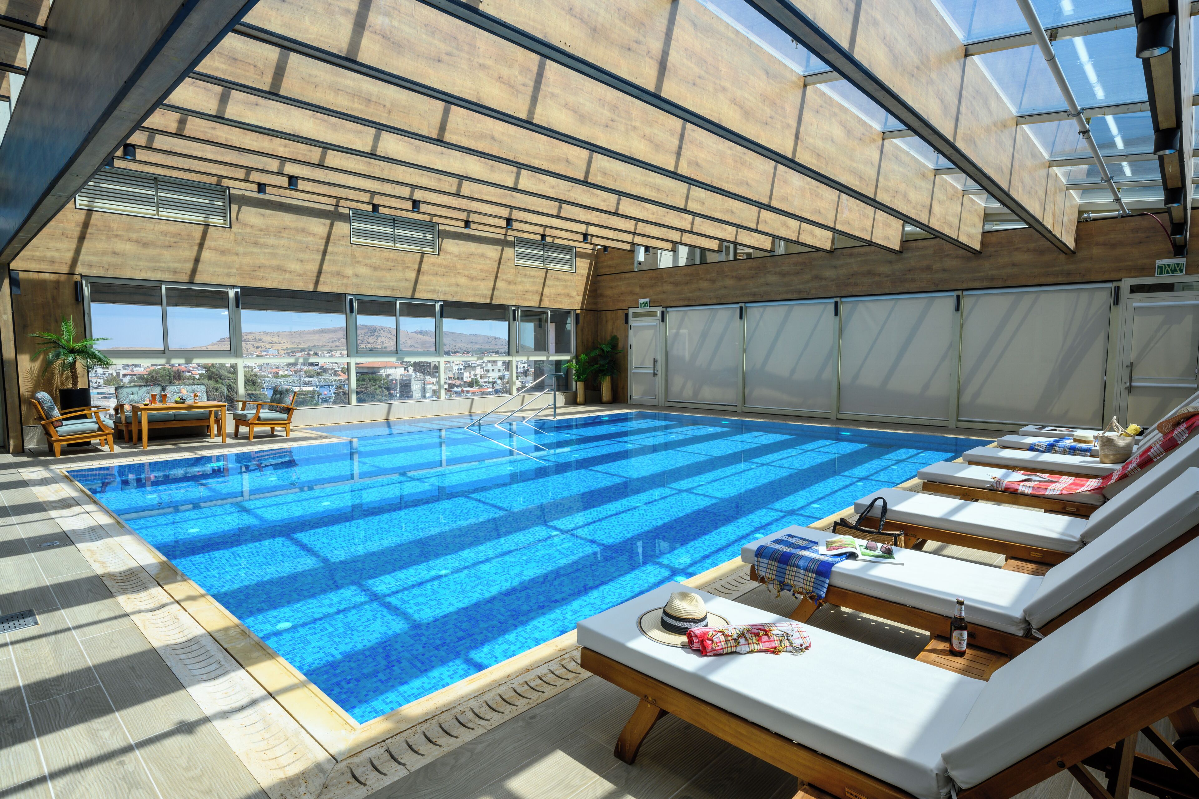 Una piscina interior moderna con agua azul cristalina, tumbonas de madera y vistas a la ciudad a través de amplias ventanas.