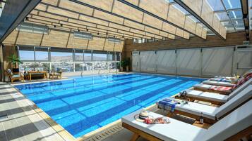 Una piscina interior moderna con agua azul cristalina, tumbonas de madera y vistas a la ciudad a través de amplias ventanas.