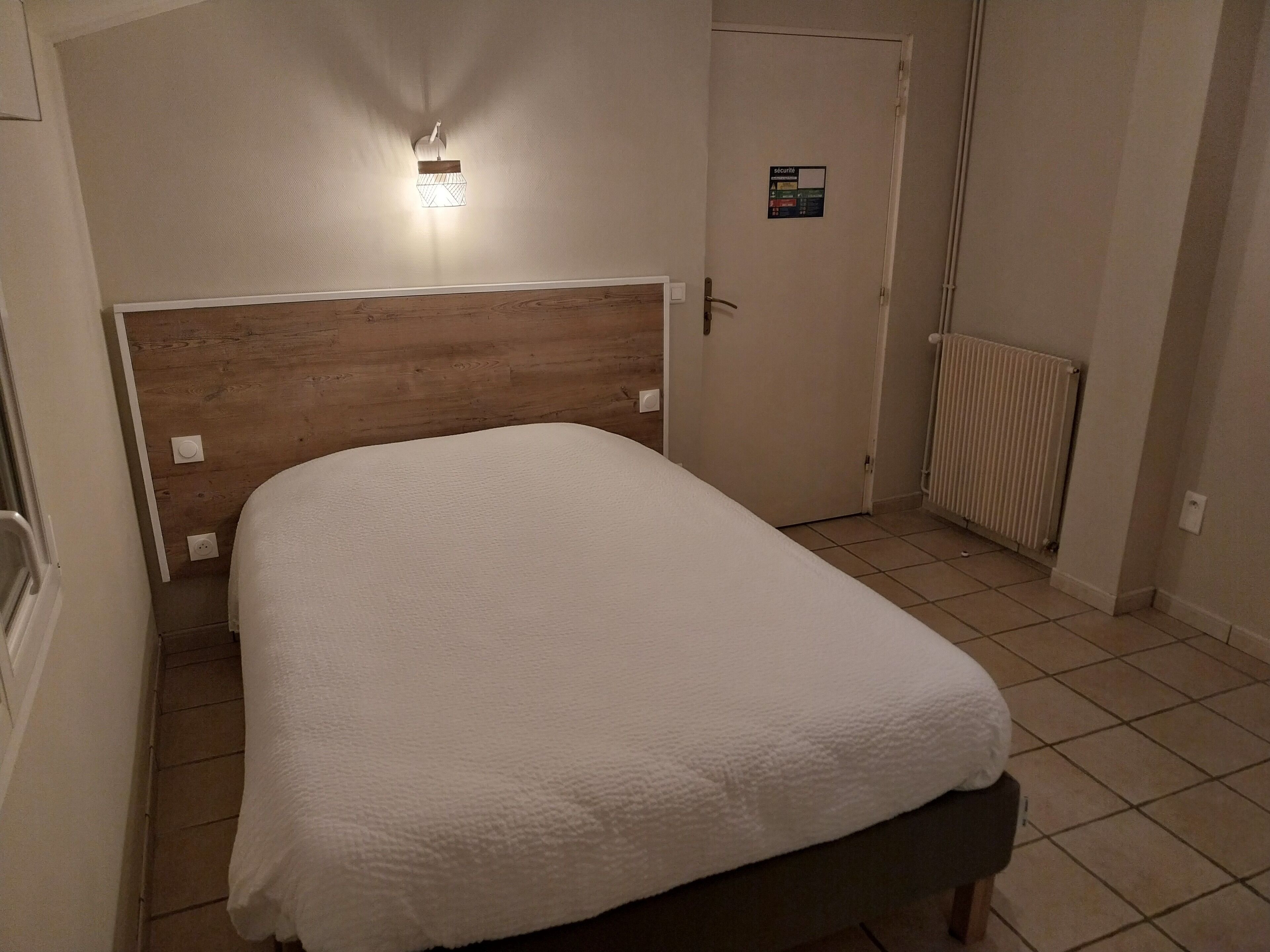 A bedroom with a wooden headboard, a white bedspread, a radiator, and a door with a security sign.