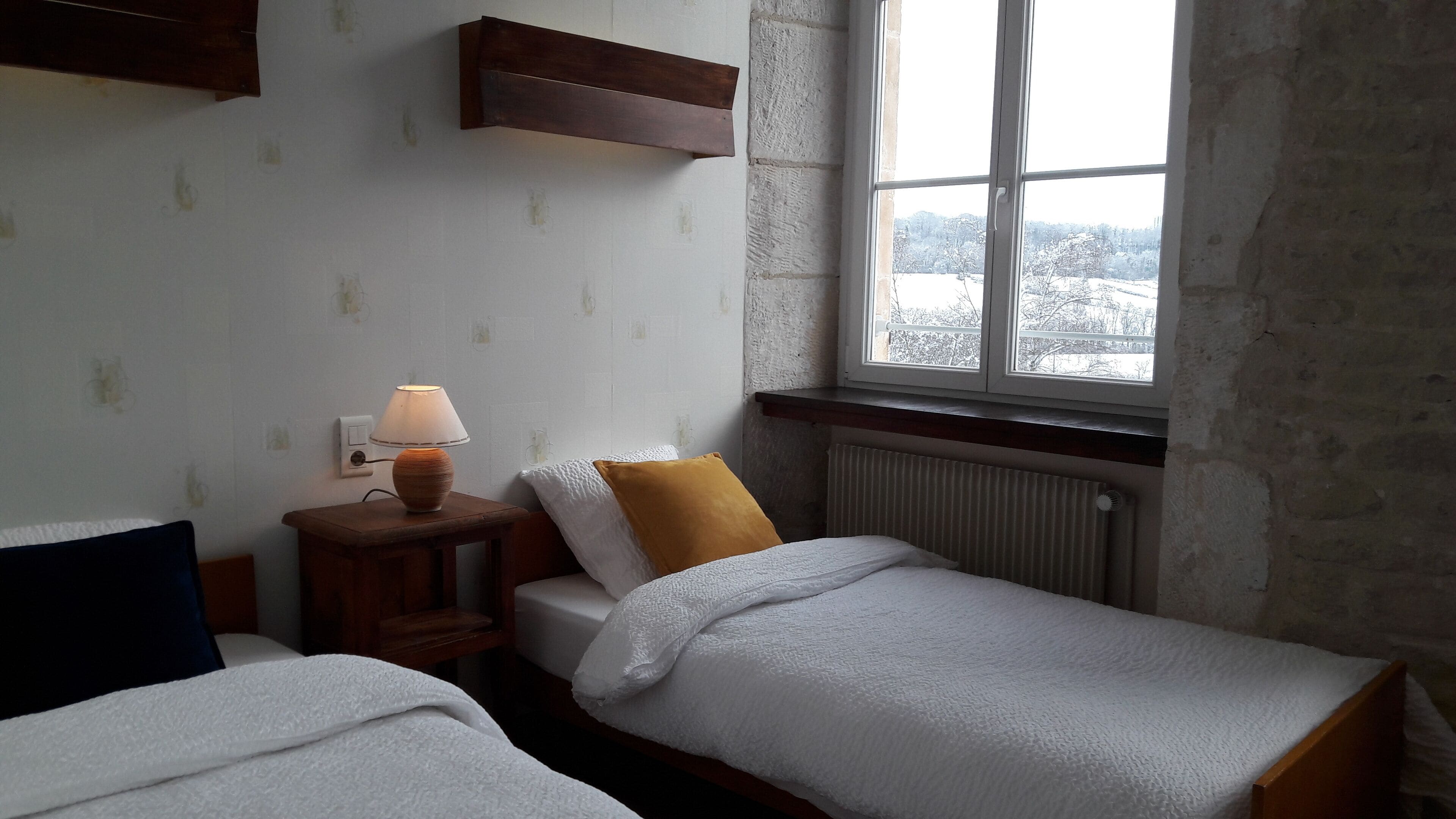 A bedroom with two single beds, a bedside table with a lamp, a window with a view of trees, and a wooden shelf above the bed.