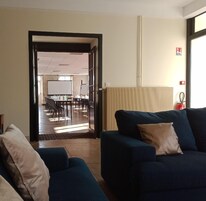 Una sala de estar moderna con un sofá azul oscuro y un cojín color beige, suelo de madera, y vistas a una sala de conferencias con una pizarra y sillas.
