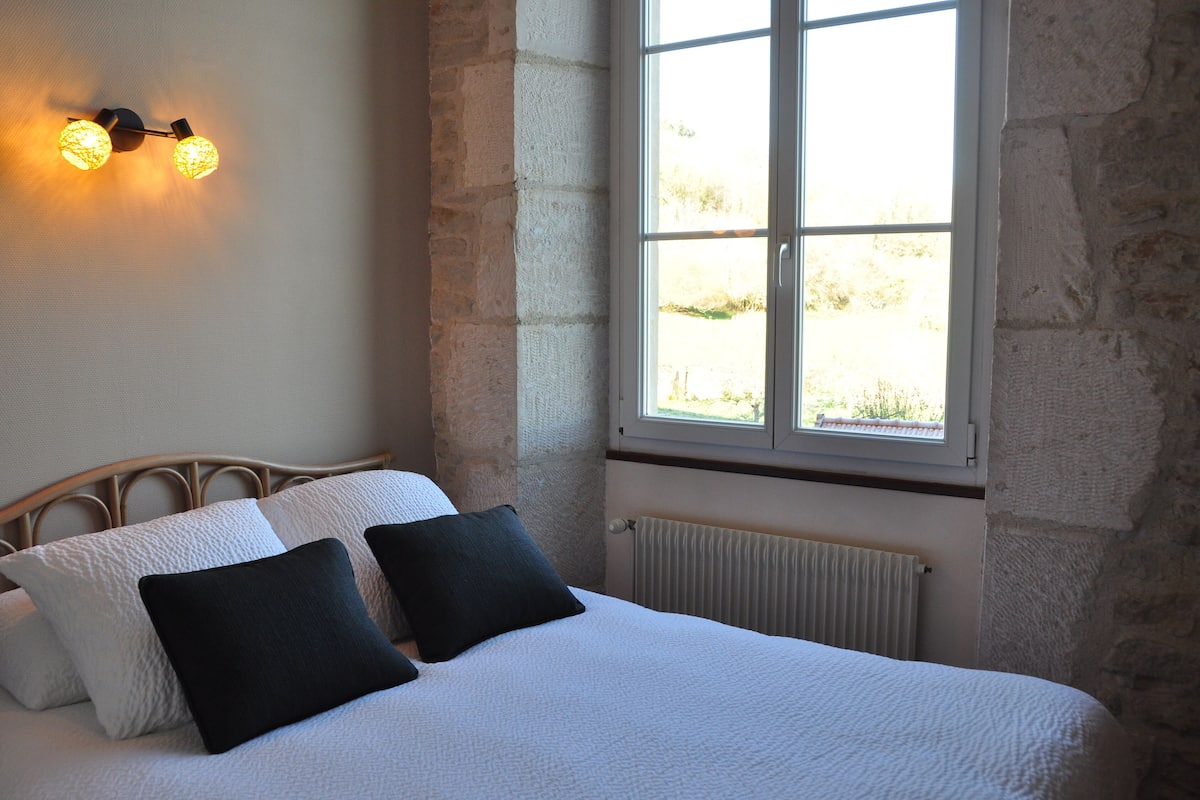 A bed with white bedding and black pillows, a stone wall, a window with a view, and a wall-mounted light fixture.