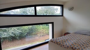 Une chambre à coucher comprenant un lit, une table de chevet et deux grandes fenêtres donnant sur un paysage verdoyant.