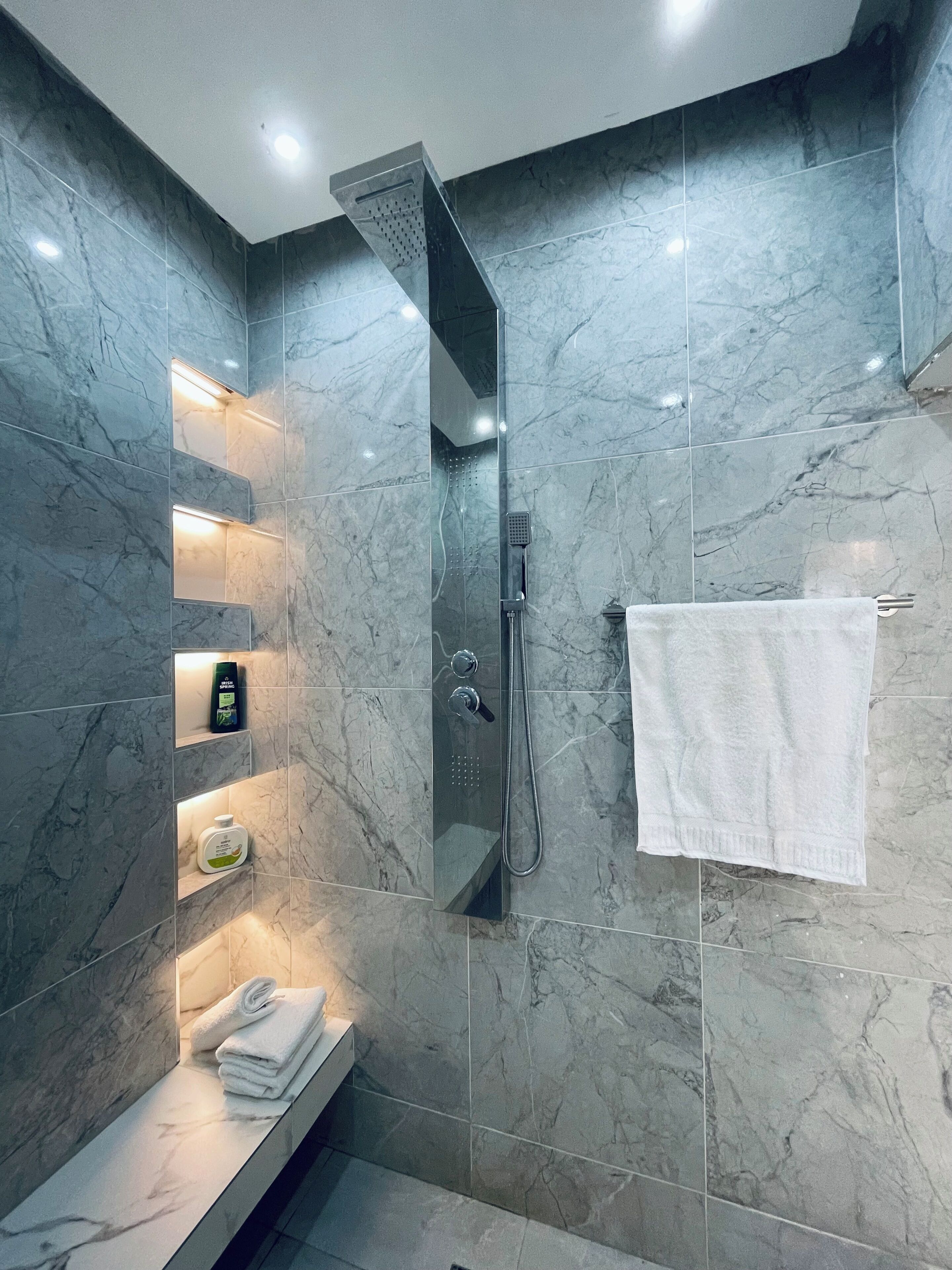 A modern bathroom with a glass-enclosed shower, marble walls, and built-in shelves with toiletries.