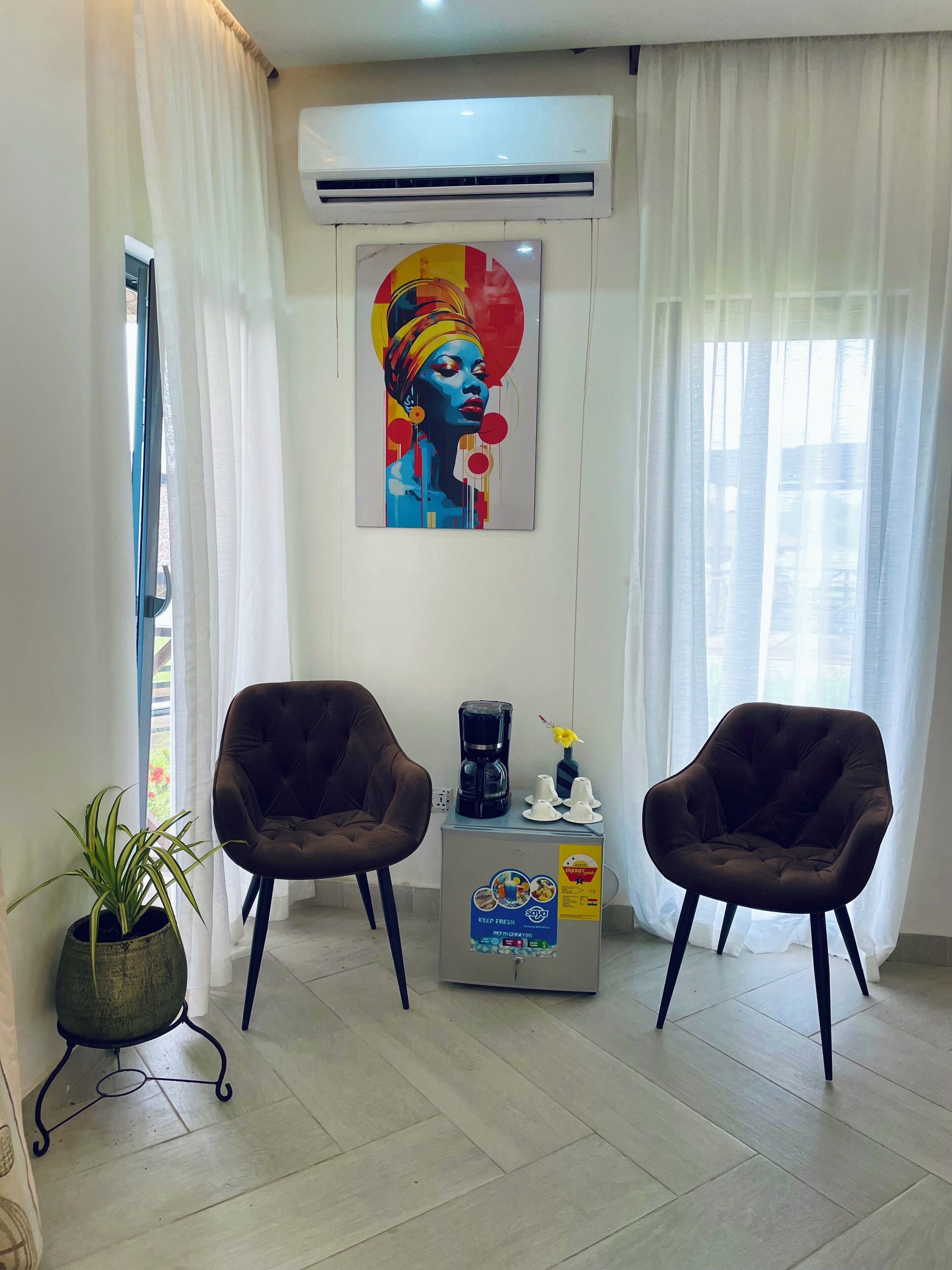 A room with two dark brown chairs, a small table with a coffee maker and cups, a potted plant, and a colorful artwork on the wall.