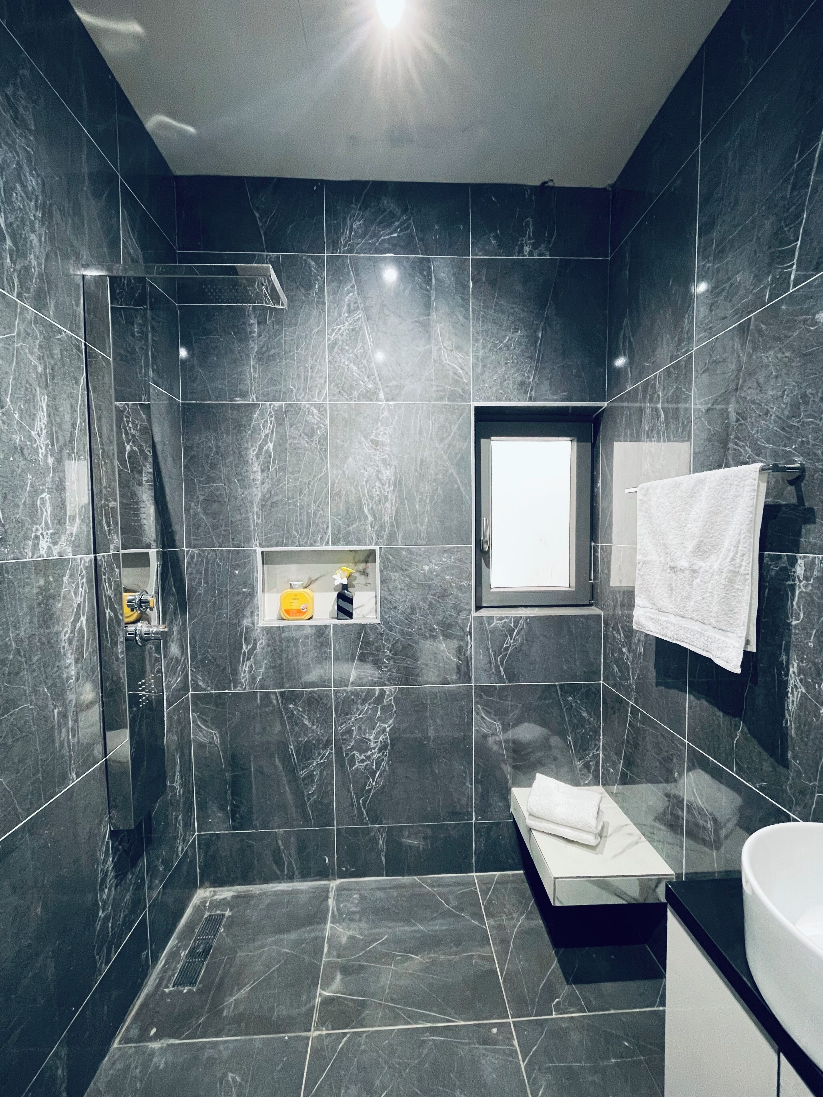 Un baño moderno con baldosas de mármol oscuro, un ventanal integrado, un perchero y una pequeña estantería con artículos de aseo.