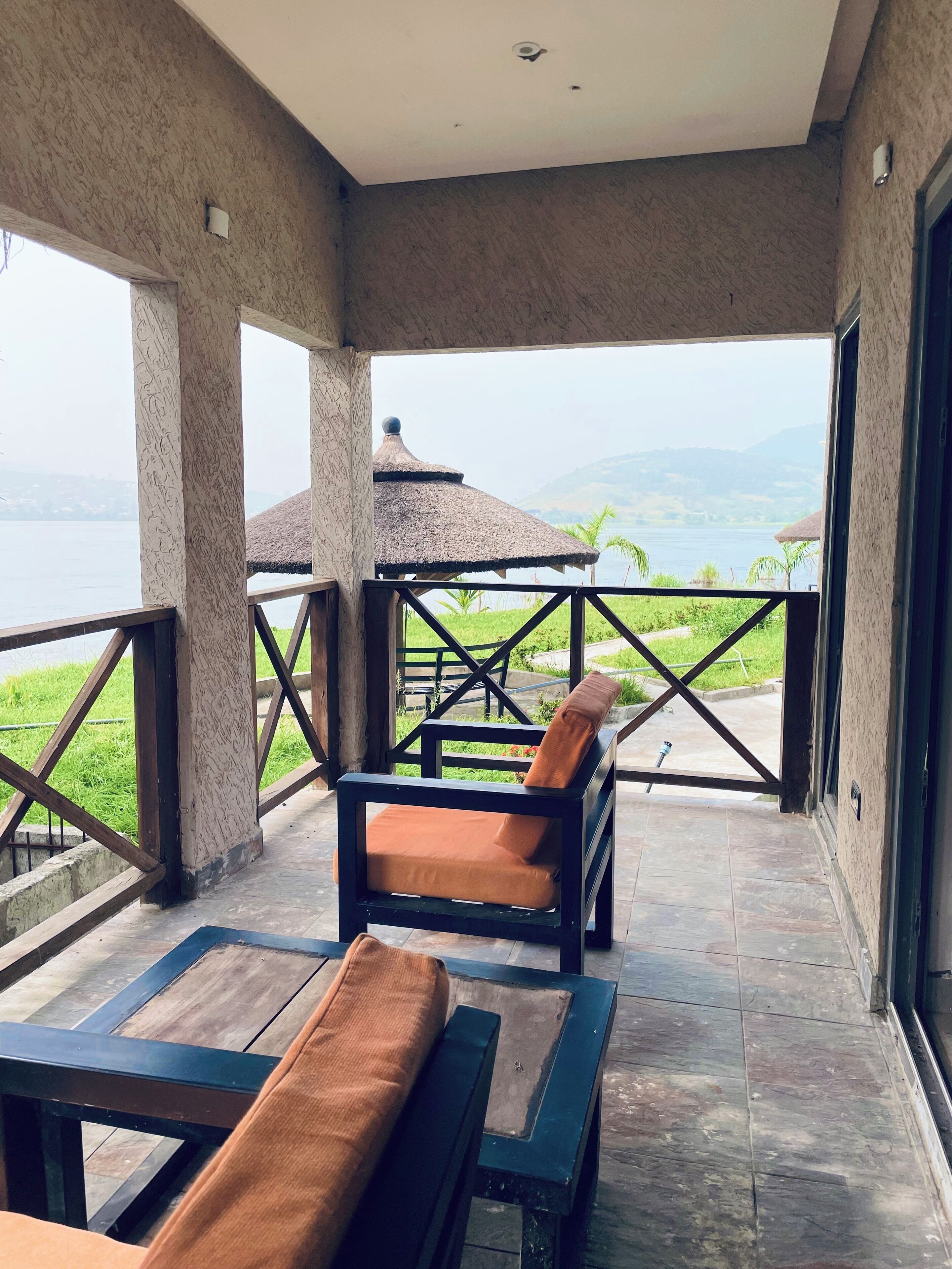 Een balkon met uitzicht op een meer en bergen, een gebouw met rieten dak en een zithoek met kussens.