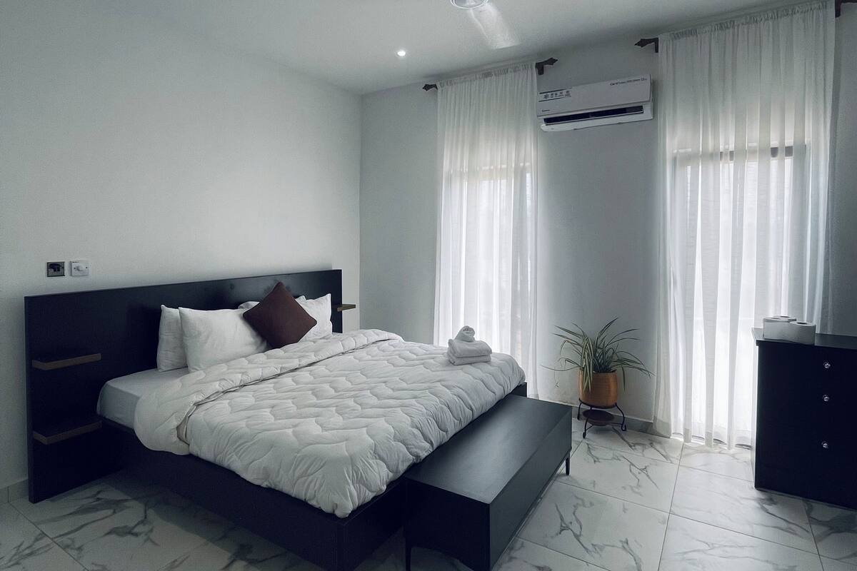 A modern bedroom with a large bed, a bedside table, a bench, and an air conditioning unit.
