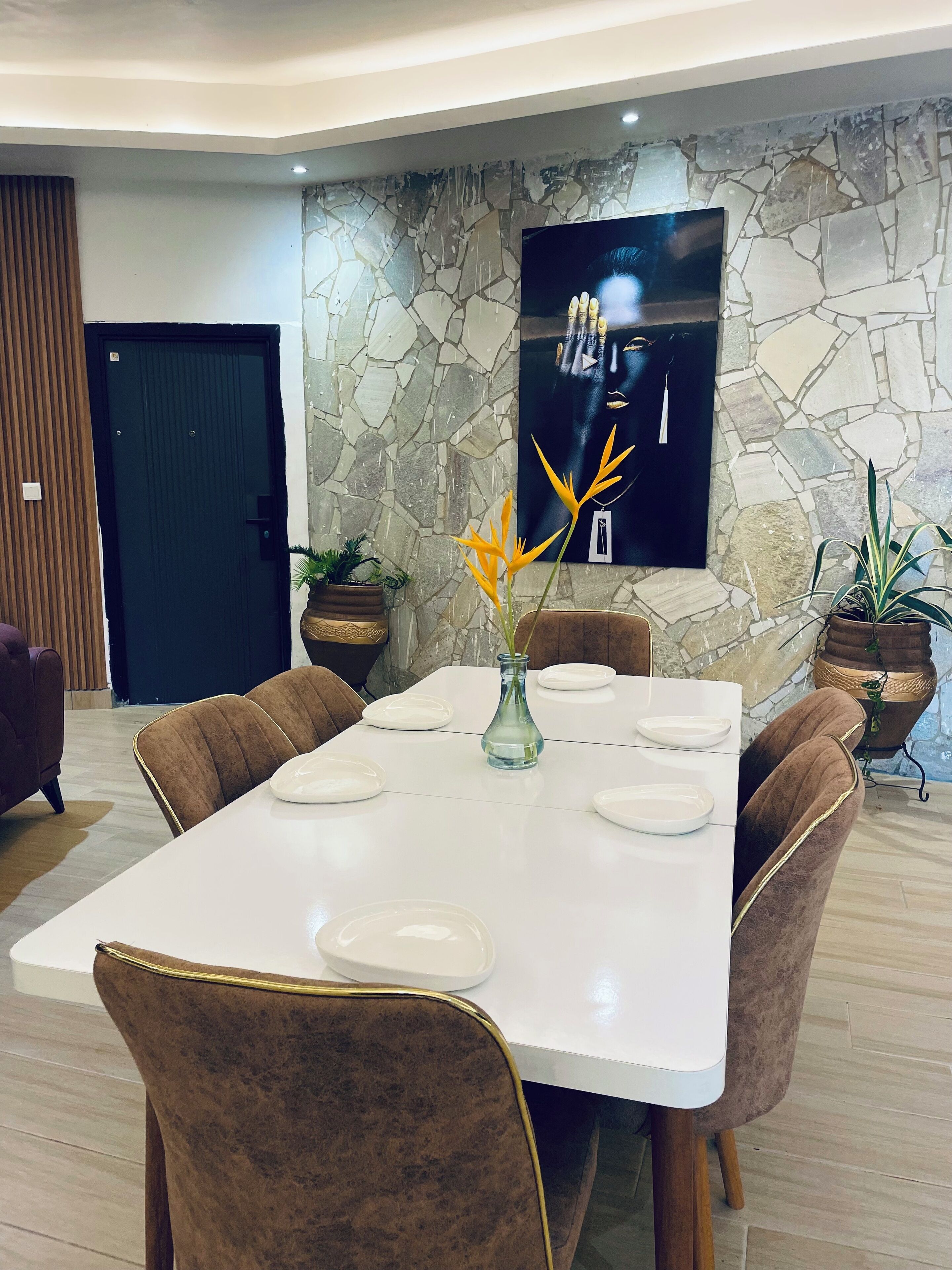 Un comedor con una mesa blanca dispuesta para seis personas, rodeado de sillas tapizadas en marrón. Hay plantas en macetas sobre la mesa y una pared de piedra con un cuadro colgado.