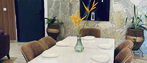 Un comedor con una mesa blanca dispuesta para seis personas, rodeado de sillas tapizadas en marrón. Hay plantas en macetas sobre la mesa y una pared de piedra con un cuadro colgado.