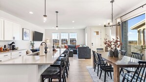 A modern kitchen with a central island, dining area with a wooden table and chairs, and a living area with a sofa and chairs. - Antler's Peak | Bridger Views & Close to Airport (Belgrade)