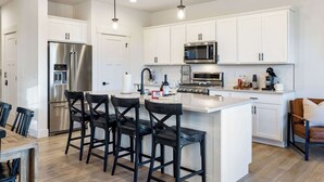 A modern kitchen with white cabinetry, stainless steel appliances, and a central island with bar stools. - Antler's Peak | Bridger Views & Close to Airport (Belgrade)