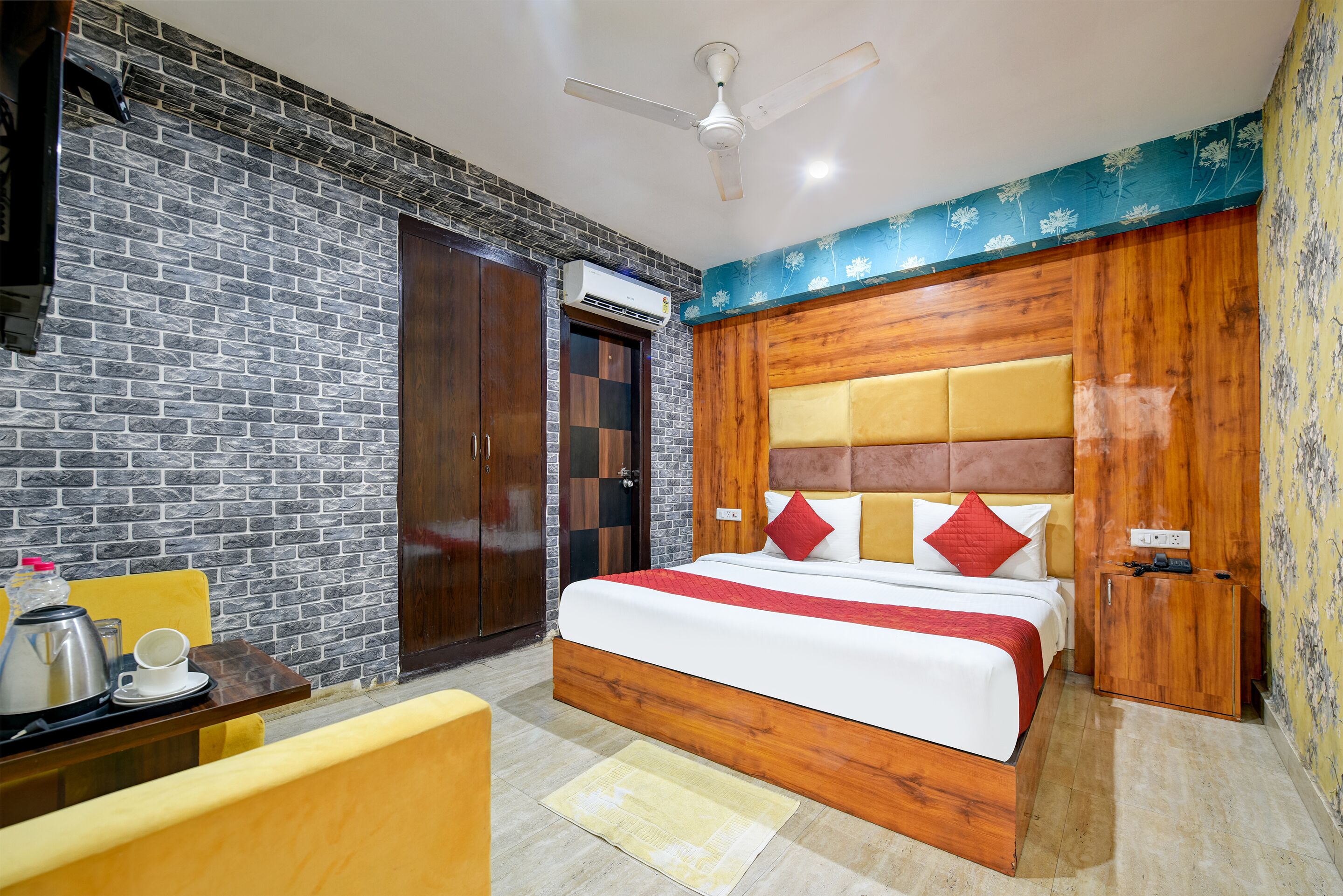 A hotel room with a brick-patterned wall, a wooden headboard, a bed with red and white bedding, a small table with a teapot and cup, and a ceiling fan.