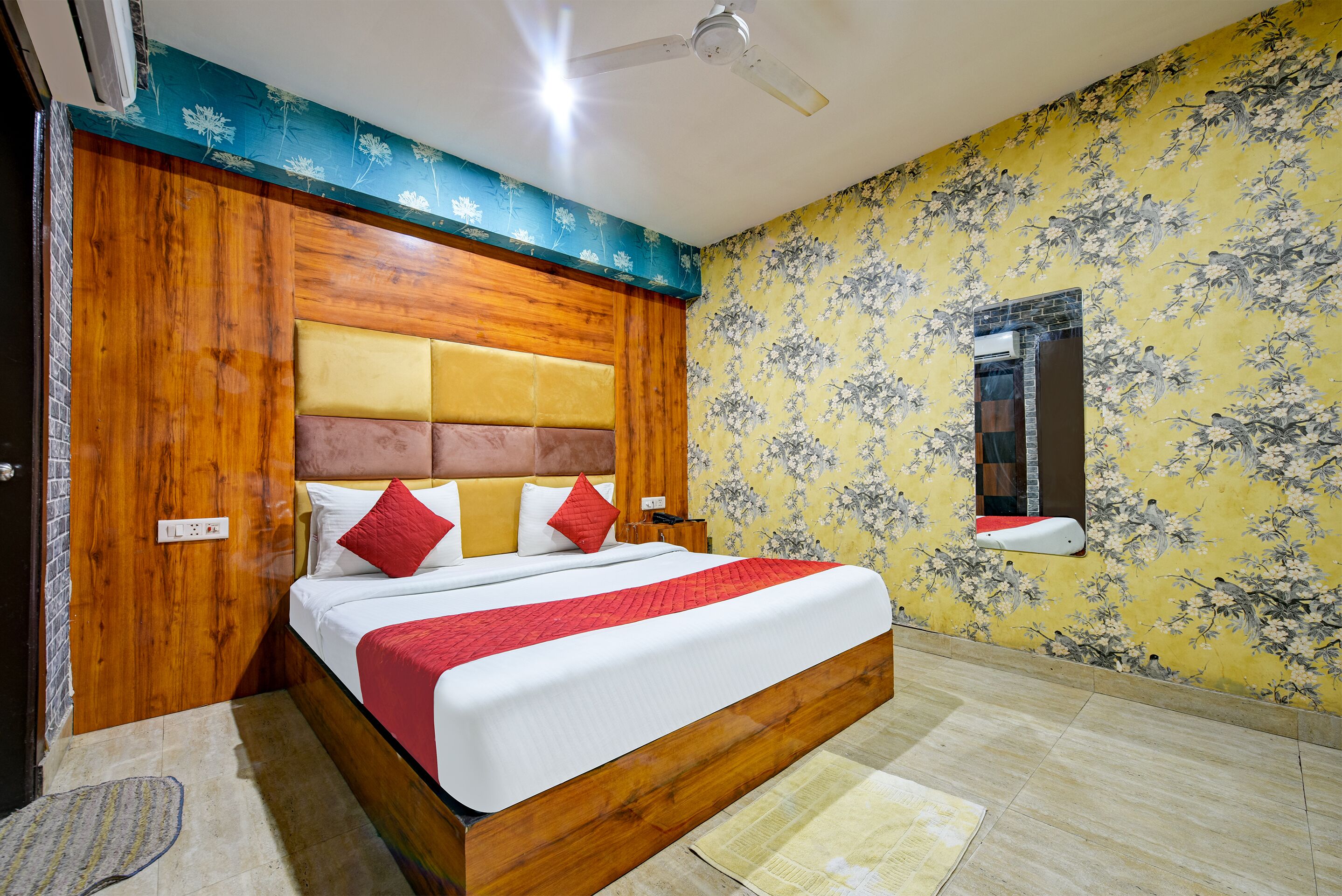 Quarto com cabeceira de madeira, cama com roupa de cama vermelha e branca, ventilador de teto e papel de parede floral.