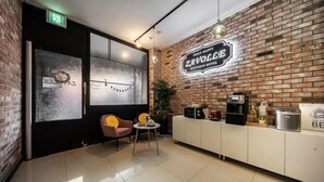A cozy hotel lobby with a brick wall, a large window with frosted glass, a seating area with a chair and table, and a reception desk with a coffee machine. - Zavolle Hotel (Suwon)