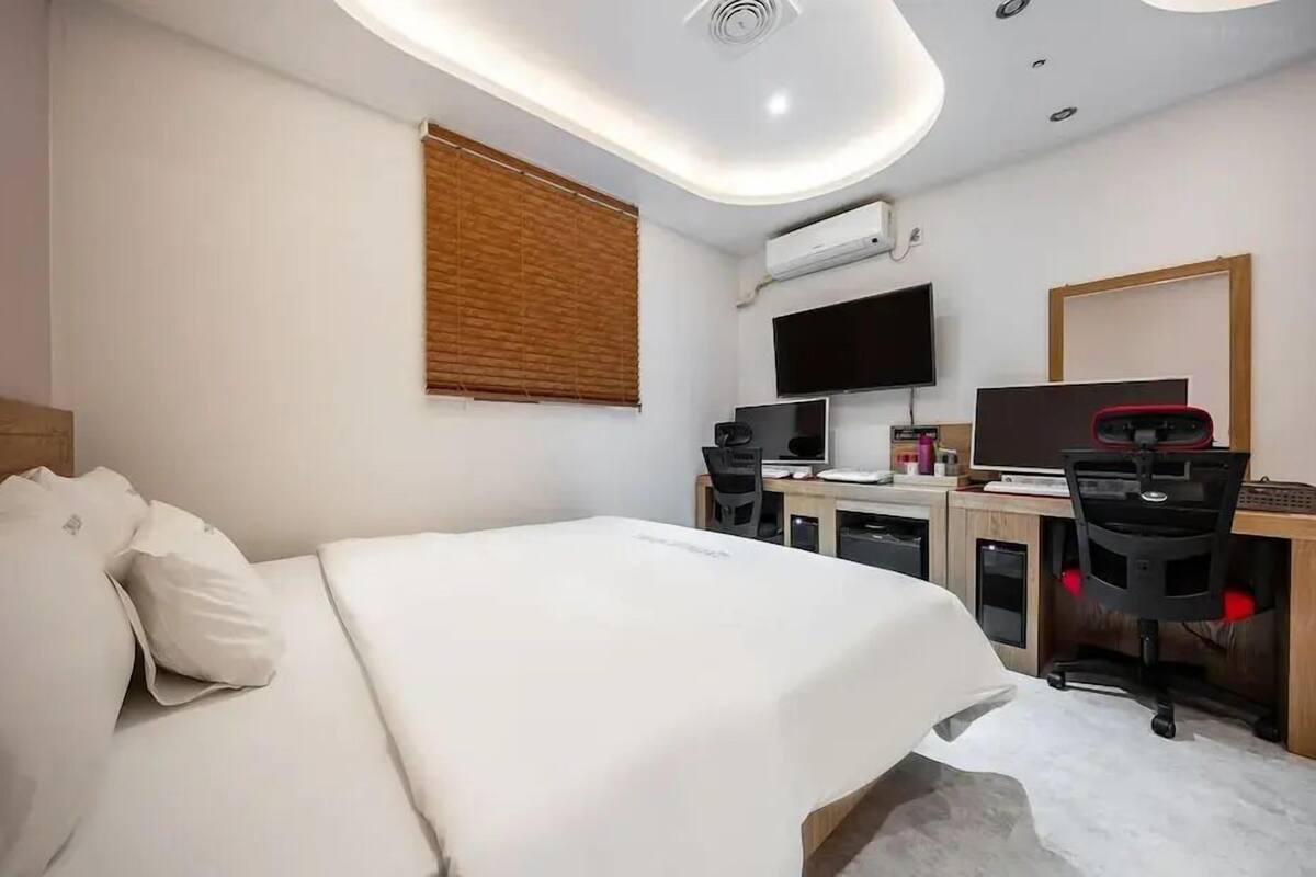 A hotel room with a bed, a desk with two monitors, a chair, and a television.